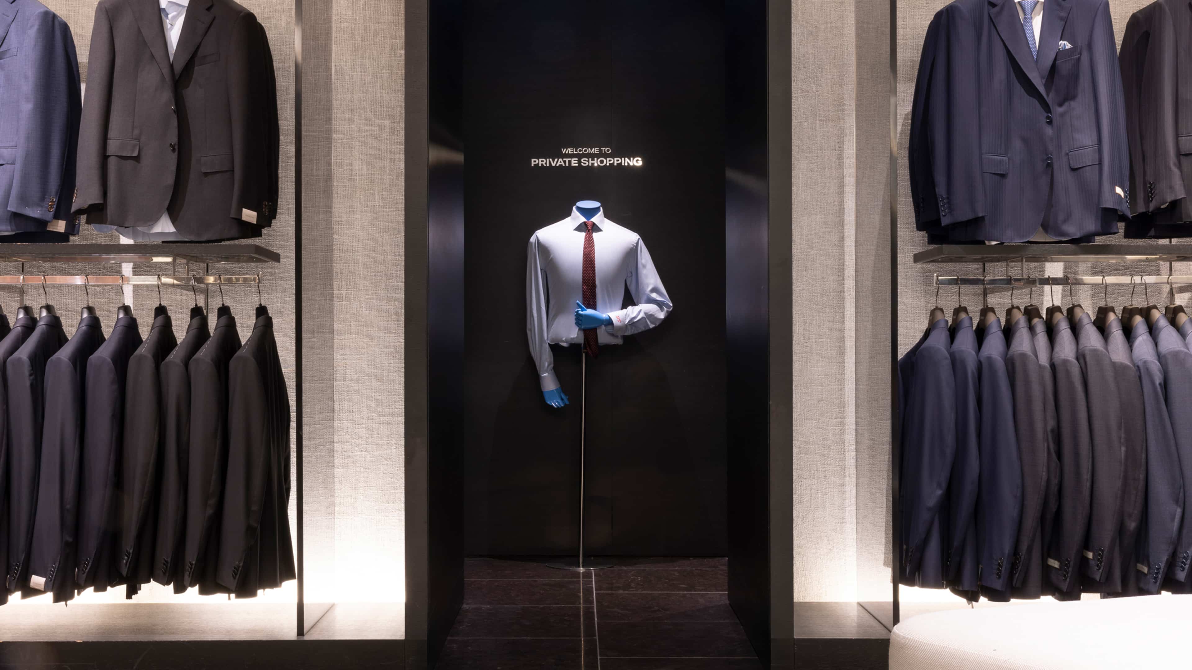 A display of suits in a clothing store with a central mannequin wearing a shirt and tie, flanked by racks of jackets and suits.