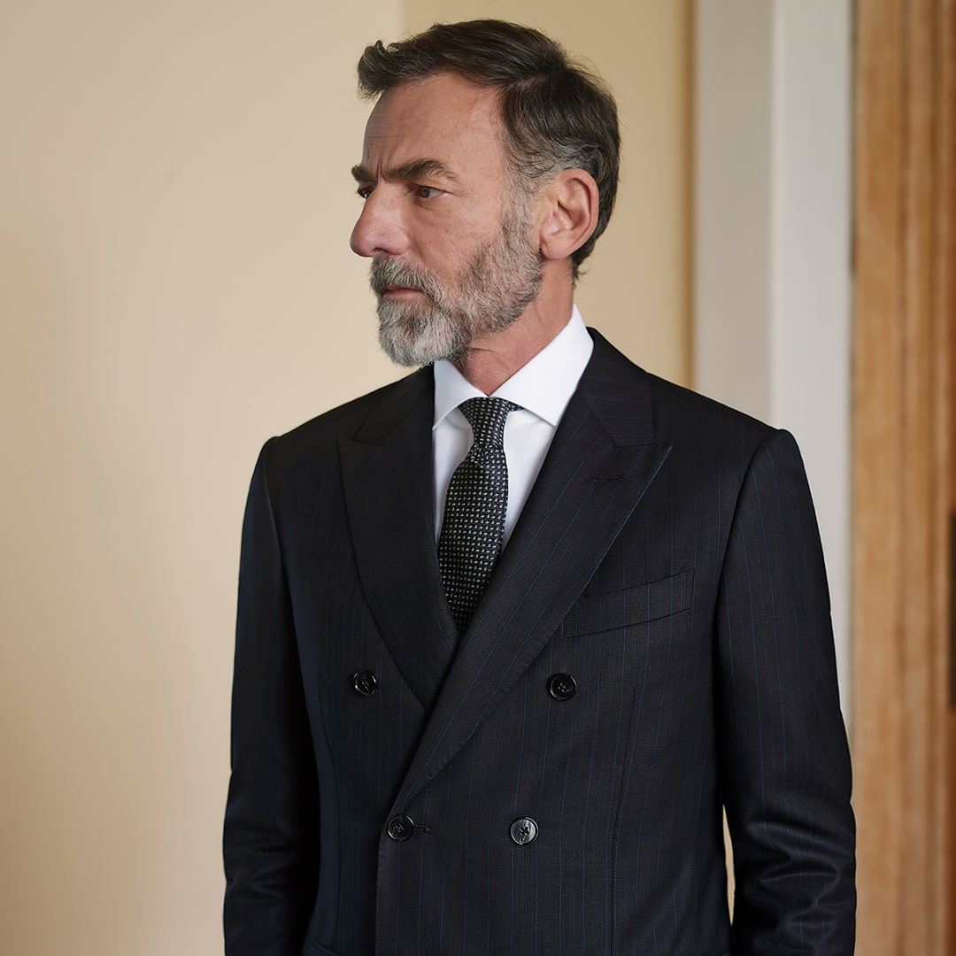 A man with a beard in a dark suit and tie stands against a light-colored wall.