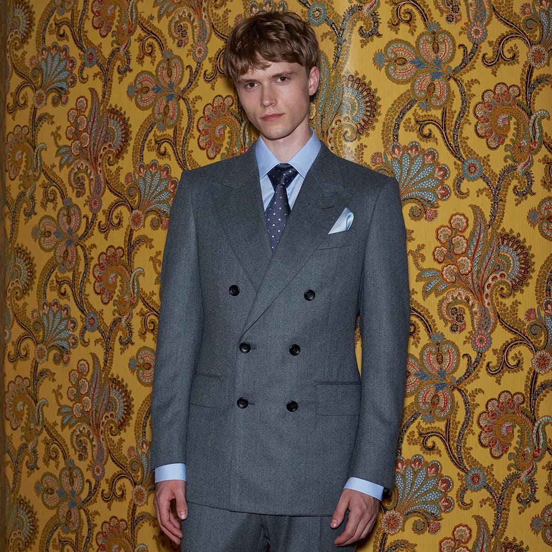 A person in a gray double-breasted suit stands against a floral patterned yellow wallpaper.