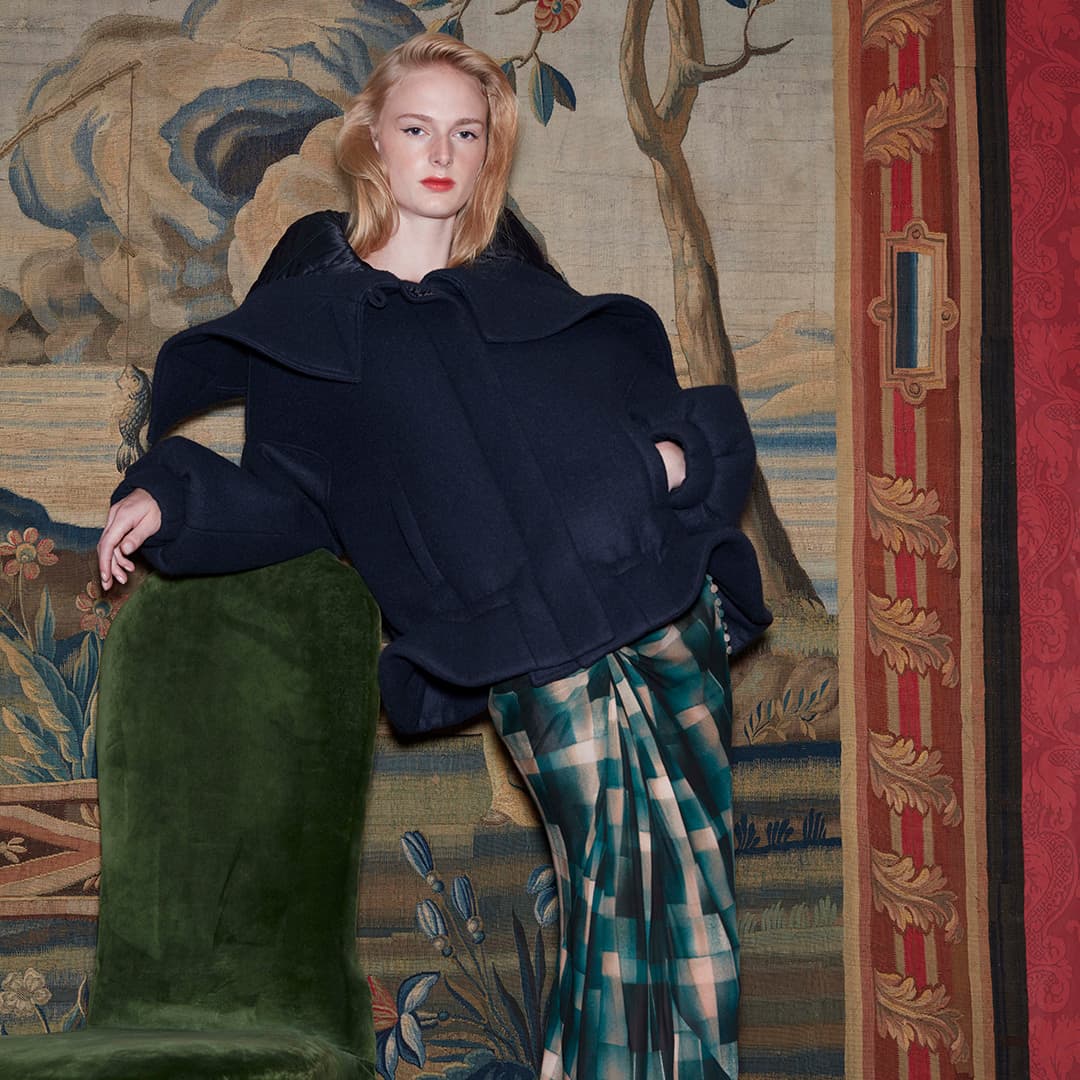 Person in a navy coat and plaid skirt stands against a tapestry backdrop beside a green velvet chair.