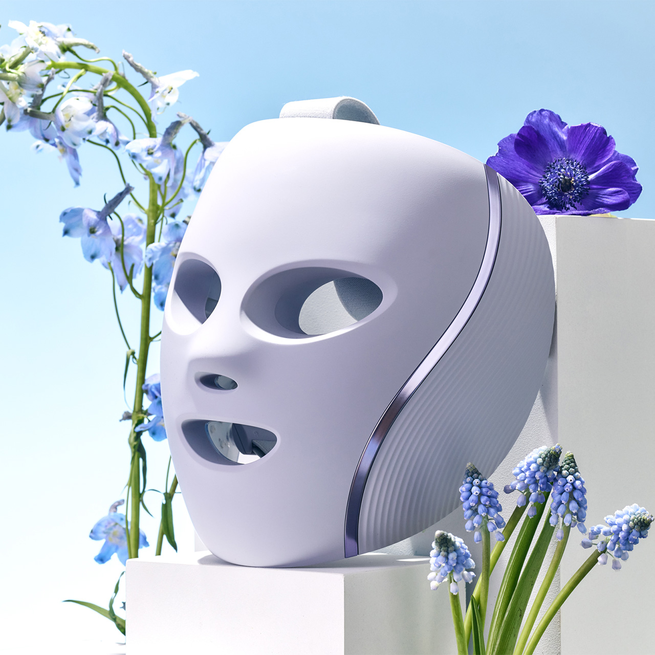 A white LED light therapy mask on a pedestal, surrounded by blue and purple flowers.