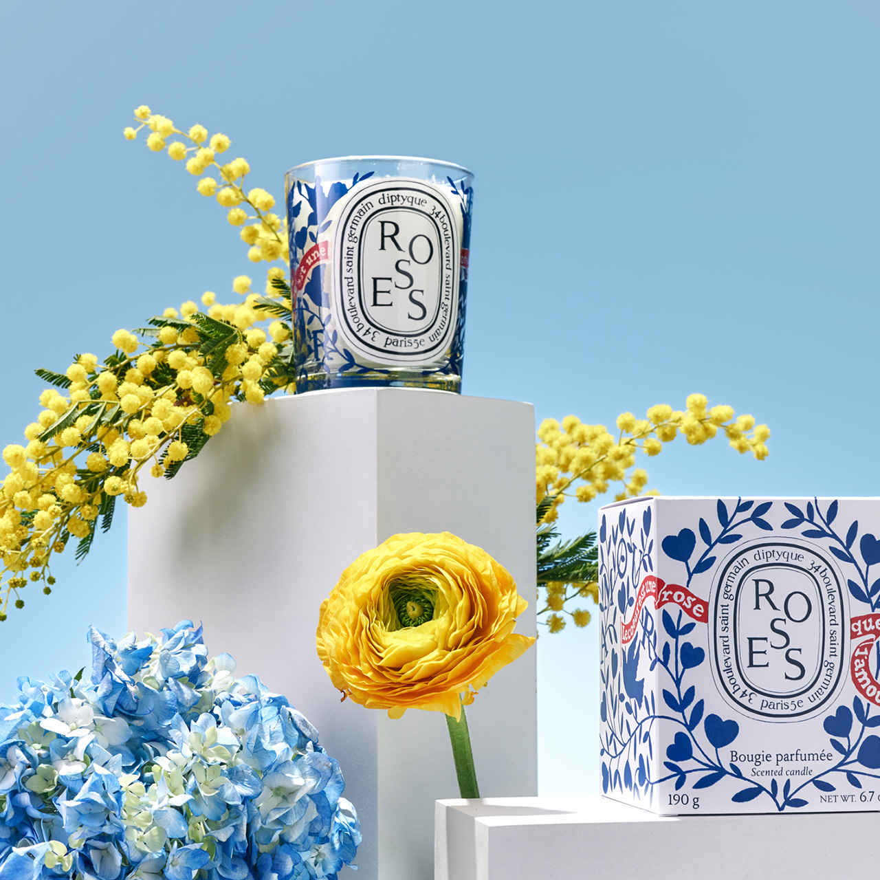 Candles with floral designs are displayed on white pedestals, surrounded by yellow and blue flowers against a clear sky background.'