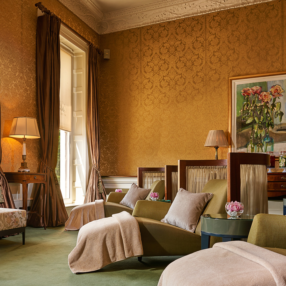 A person in a robe walks past sauna doors near an indoor pool, with warm lighting and decorative chairs.