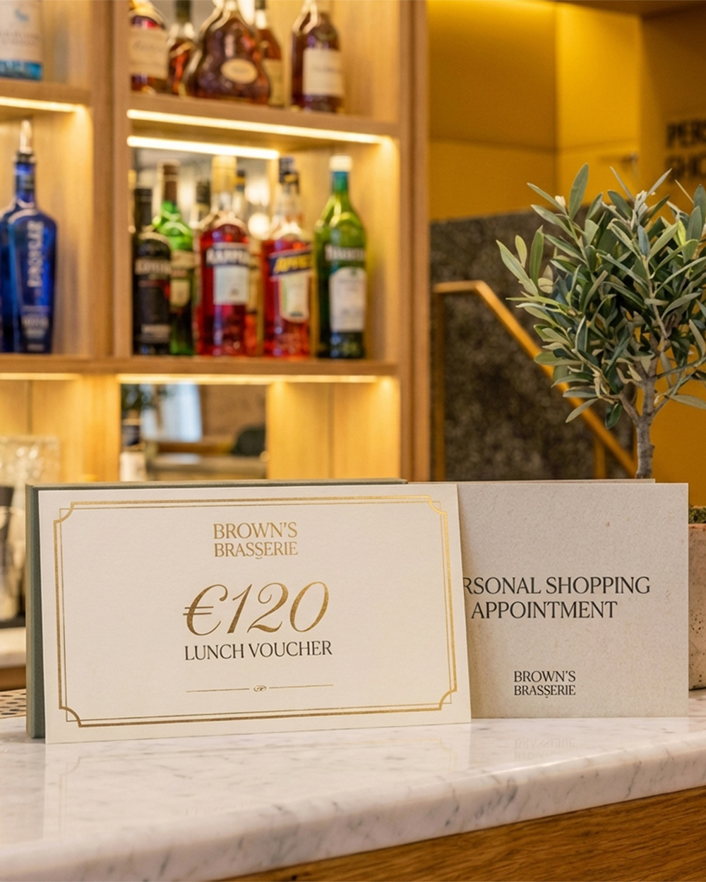 A marble counter displays a €120 lunch voucher, a personal shopping appointment card, a plant, and a glass of champagne, with a lit bar background.