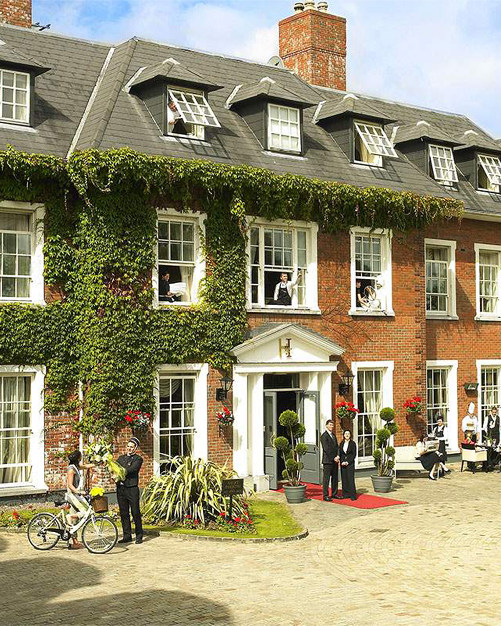 A large, ivy-covered brick building with multiple windows, people outside, and a red carpet leading to the entrance.