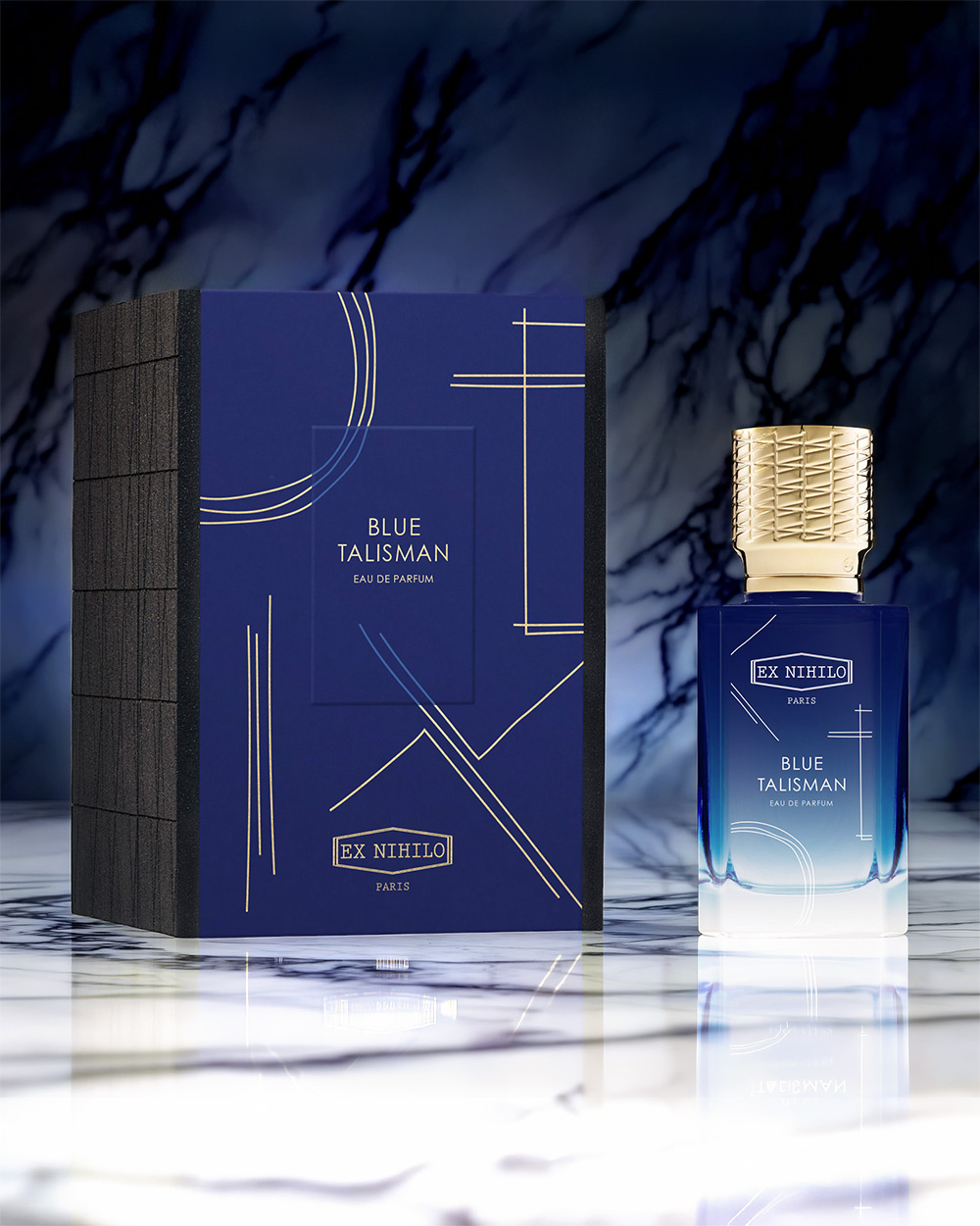 A blue and gold perfume bottle labeled 'Blue Talisman' stands next to its matching box on a marble surface.