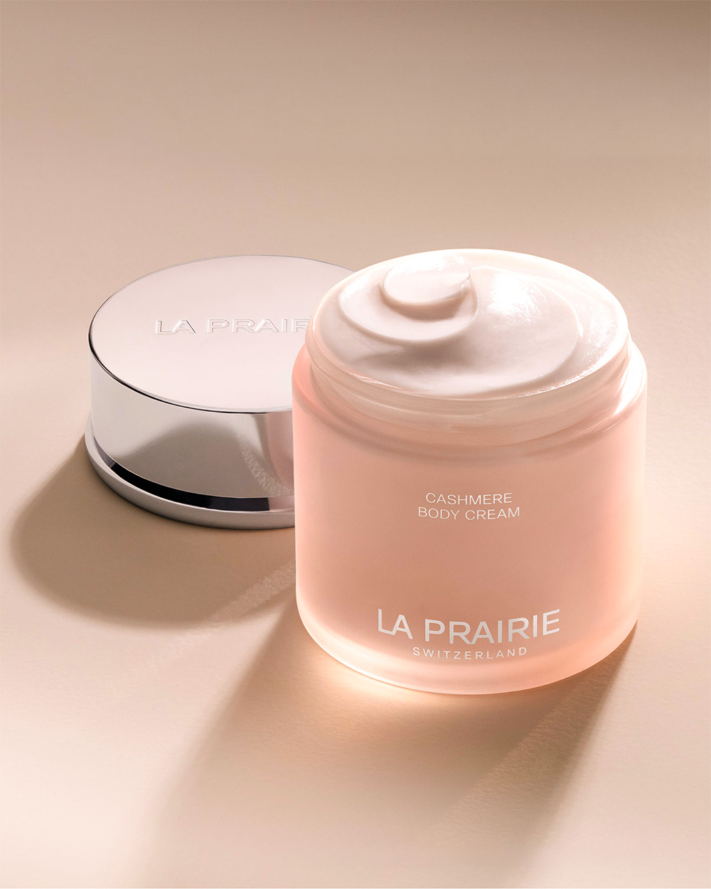A jar of La Prairie Cashmere Body Cream with its lid beside it on a beige surface.
