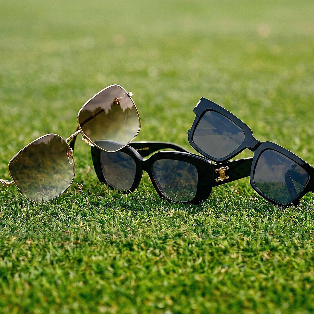 Three pairs of designer sunglasses in various styles displayed on grass in sunlight