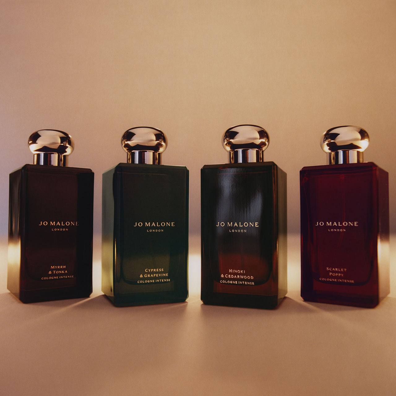 Four Jo Malone London cologne bottles with gold caps in dark glass, displaying Myrrh & Tonka, Cypress & Grapevine, Hinoki & Cedarwood, and Scarlet Poppy fragrances