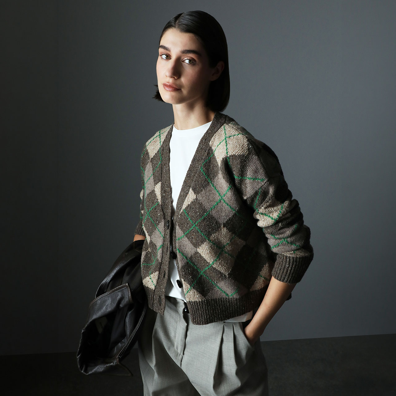 Woman wearing an argyle cardigan in grey, cream and green over a white t-shirt, holding a black leather jacket