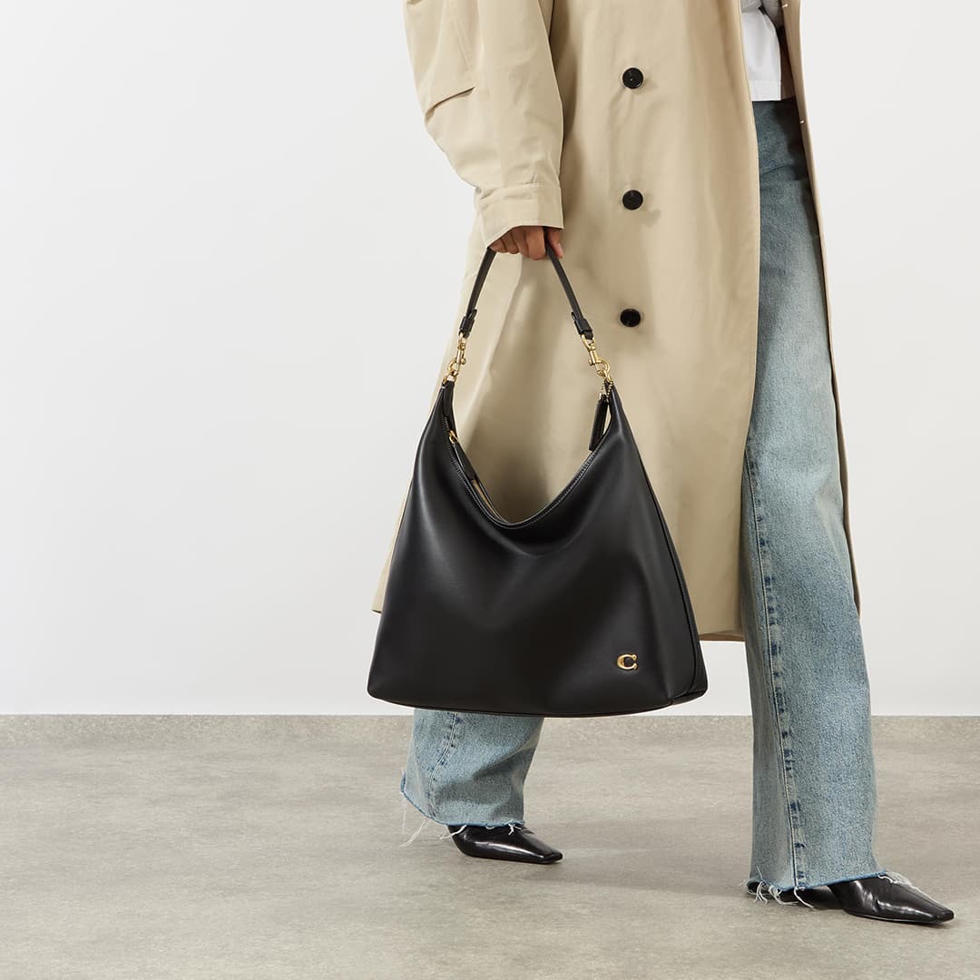 Woman holding black leather slouchy tote bag, wearing cream double-breasted coat and light blue jeans with black shoes