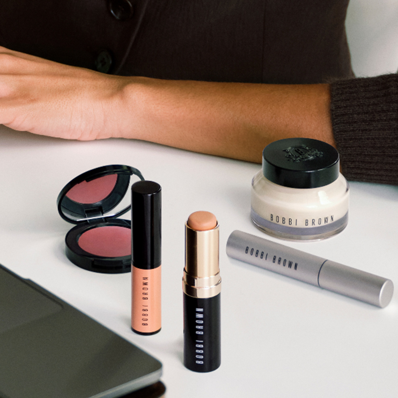 Bridal beauty products including makeup brushes, pencils, tortoiseshell hair clip, and cosmetics arranged on cream fabric