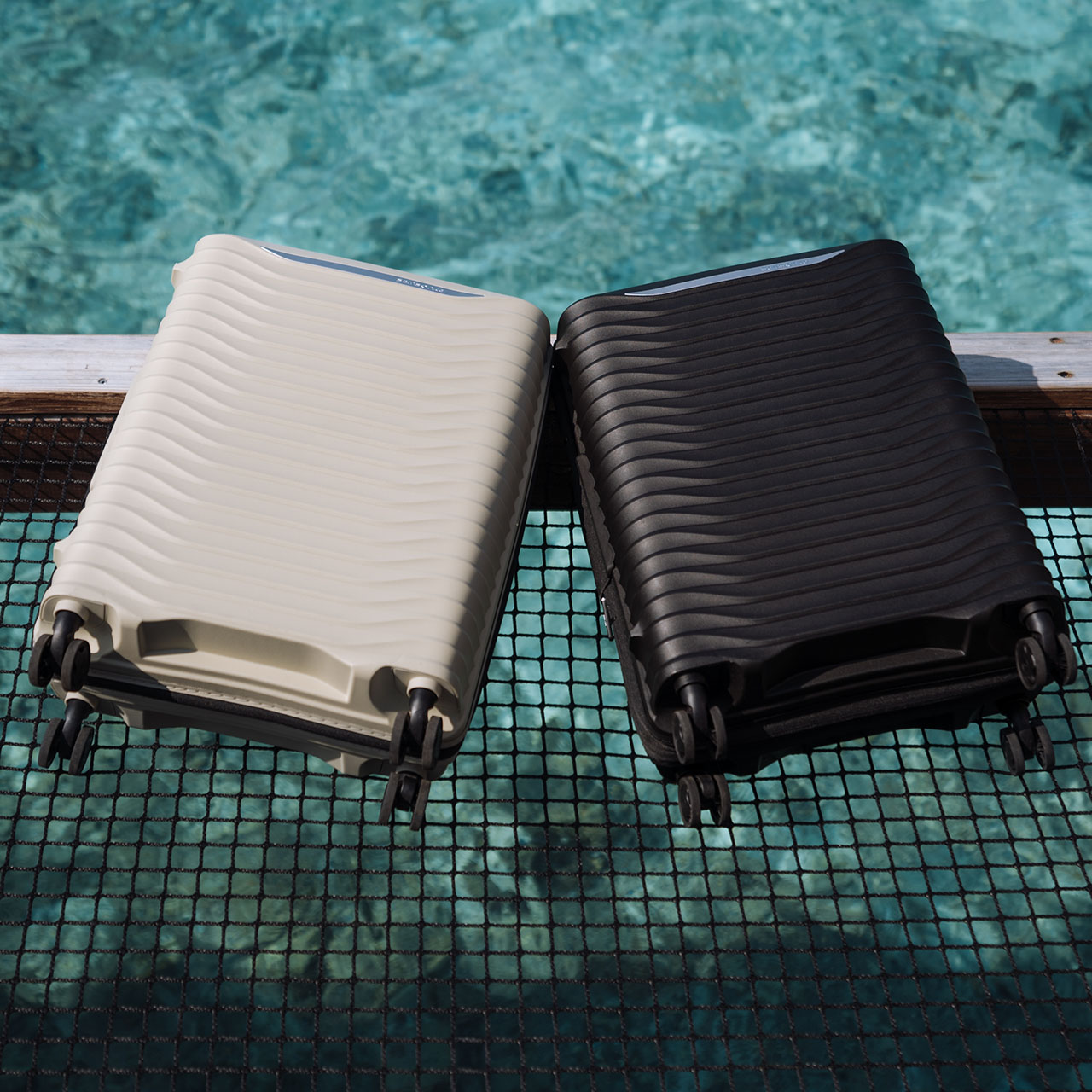 White and black hard-shell rolling suitcases positioned poolside with turquoise water and pool netting in background.