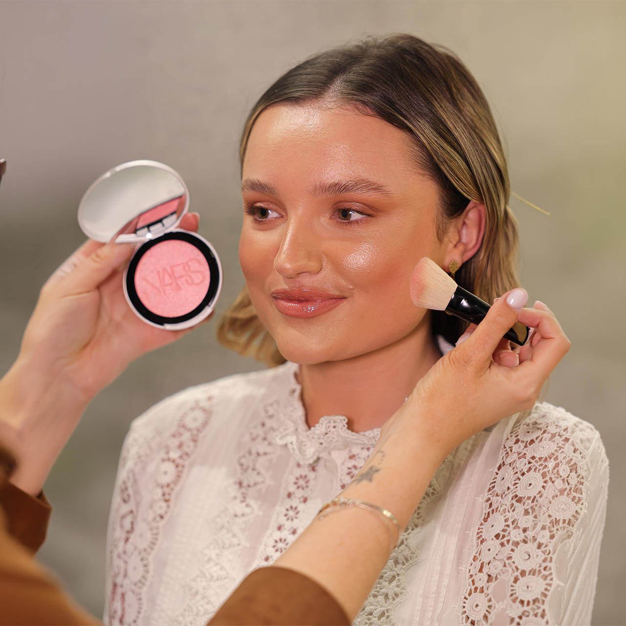 Woman applying pink blush with brush, holding open compact of pink powder with NARS branding visible
