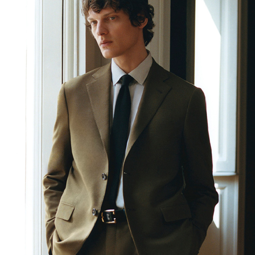 Man in a green suit and black tie stands by a window, looking away.