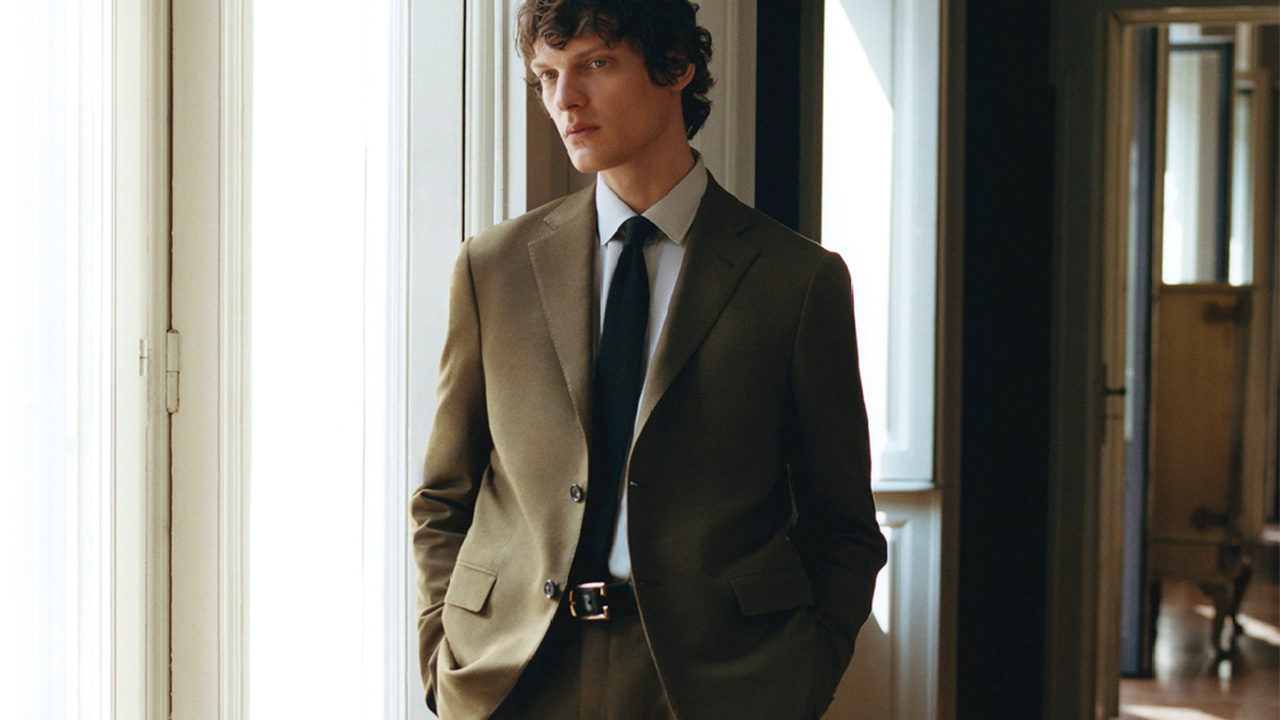 Person in a brown suit with a black tie standing near a bright window in a hallway.