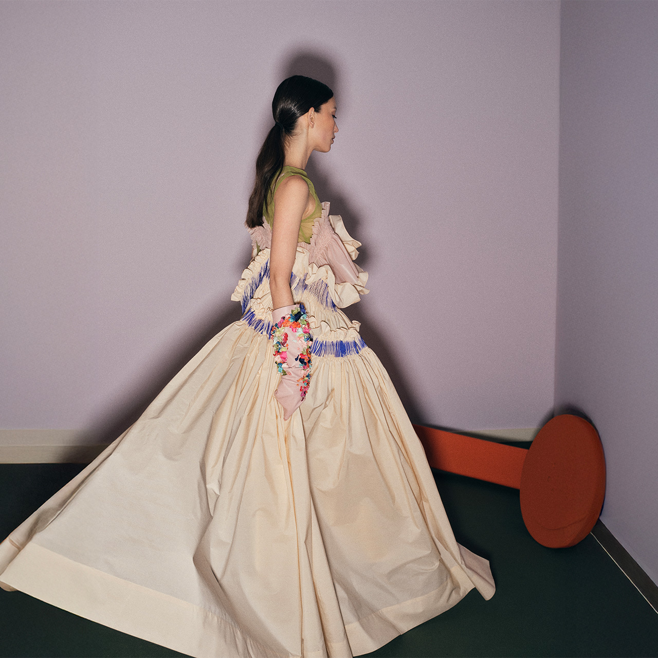 A woman in an elaborate dress with a full skirt walks in a room with pastel walls and a large orange circular object.