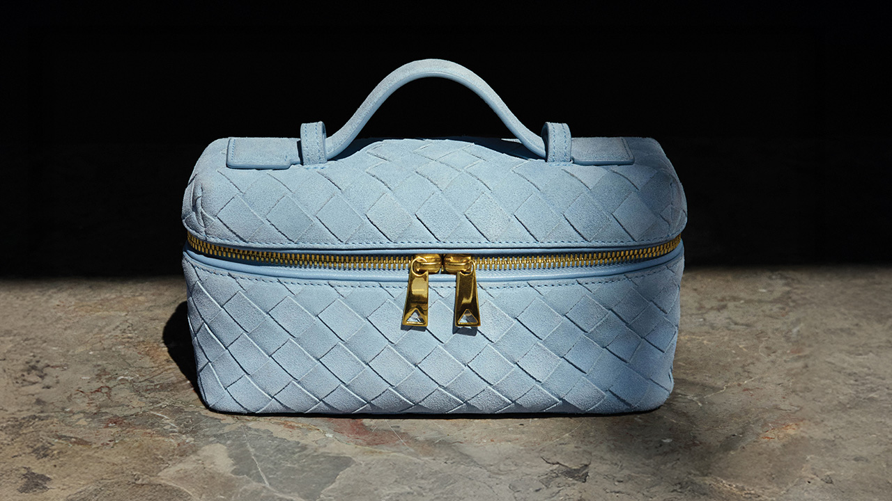 Light blue woven Bottega Veneta handbag with a gold zipper and handle, placed in shadow.