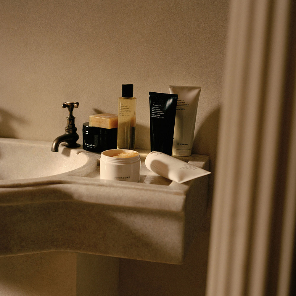Luxury bodycare products arranged on bathroom sink edge: cleansers, exfoliants, moisturisers, and soap, with brass tap visible