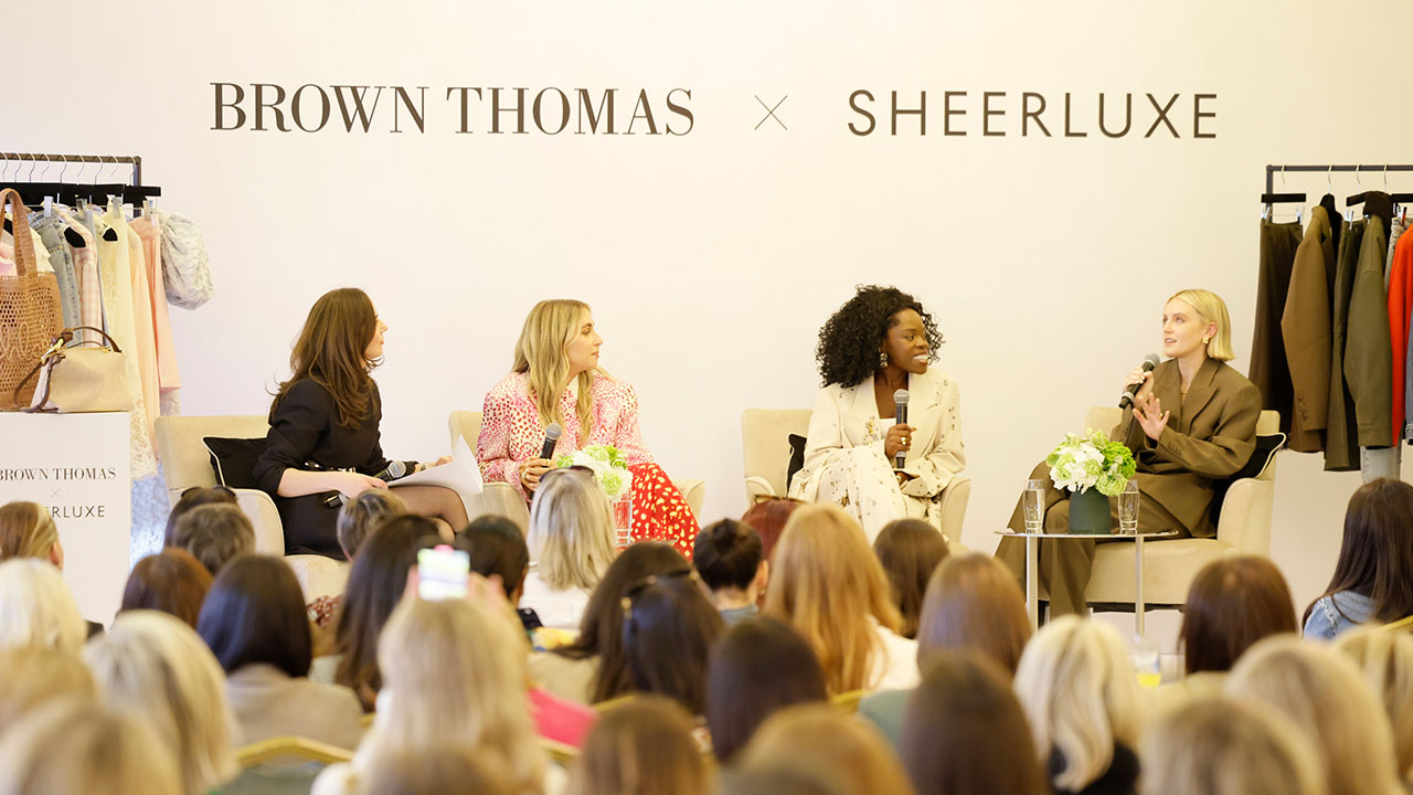 A panel of four women seated in front of an audience at a fashion event with racks of clothing in the background.