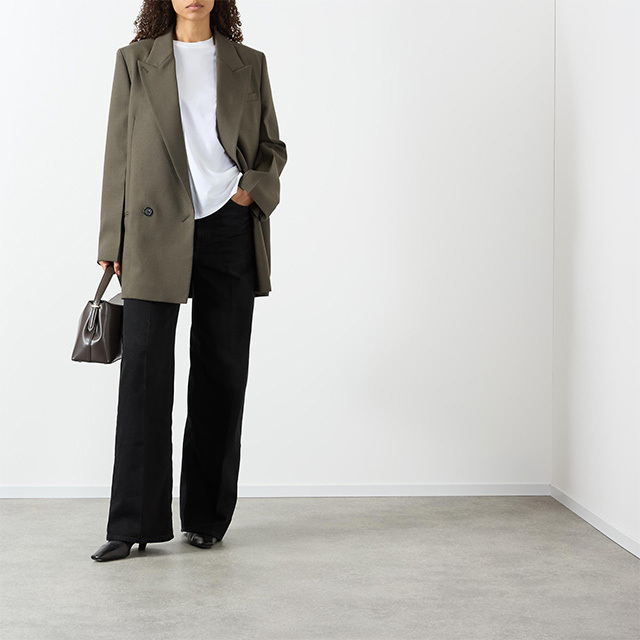 Person in oversized blazer, white shirt, and black pants holding a small handbag, standing in a minimalist room.