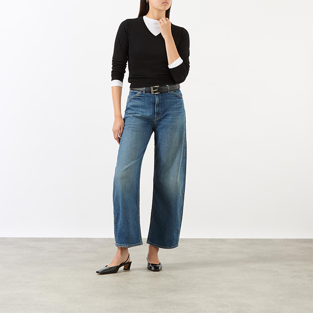 Person wearing a black sweater, white shirt, tapered jeans, and black heels, standing against a plain background.
