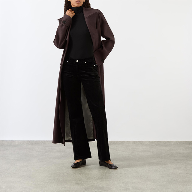 Person wearing a long brown coat, black turtleneck, black pants, and black shoes, standing on a concrete floor against a white background.