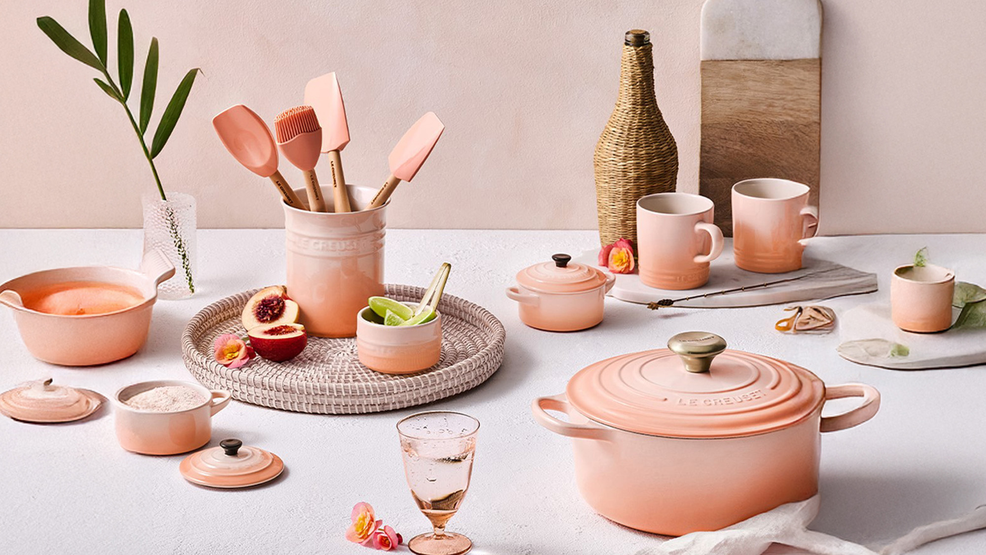 Peach-colored kitchenware set with pots, cups, utensils, and a cheese board on a light surface, accented with fruit and flowers.
