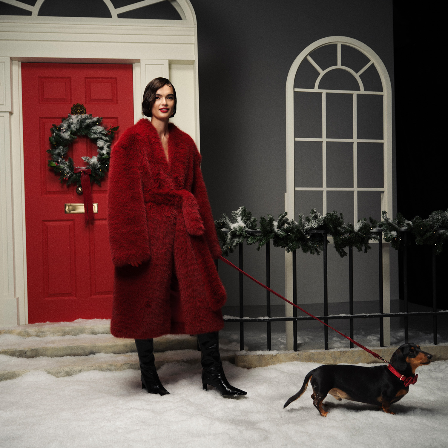 Woman in a red coat standing on snow-covered steps with a dachshund, next to a festive red door adorned with a wreath.