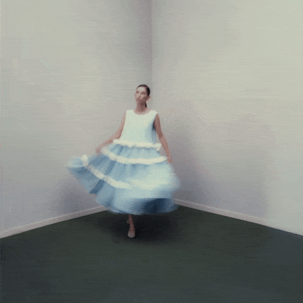 A woman in a blue and white layered dress spins in a corner of a minimalist room.