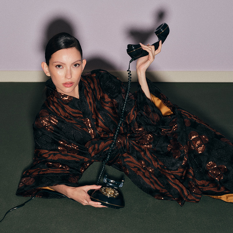Person in a patterned robe holds a vintage phone, sitting on the floor against a light purple wall.