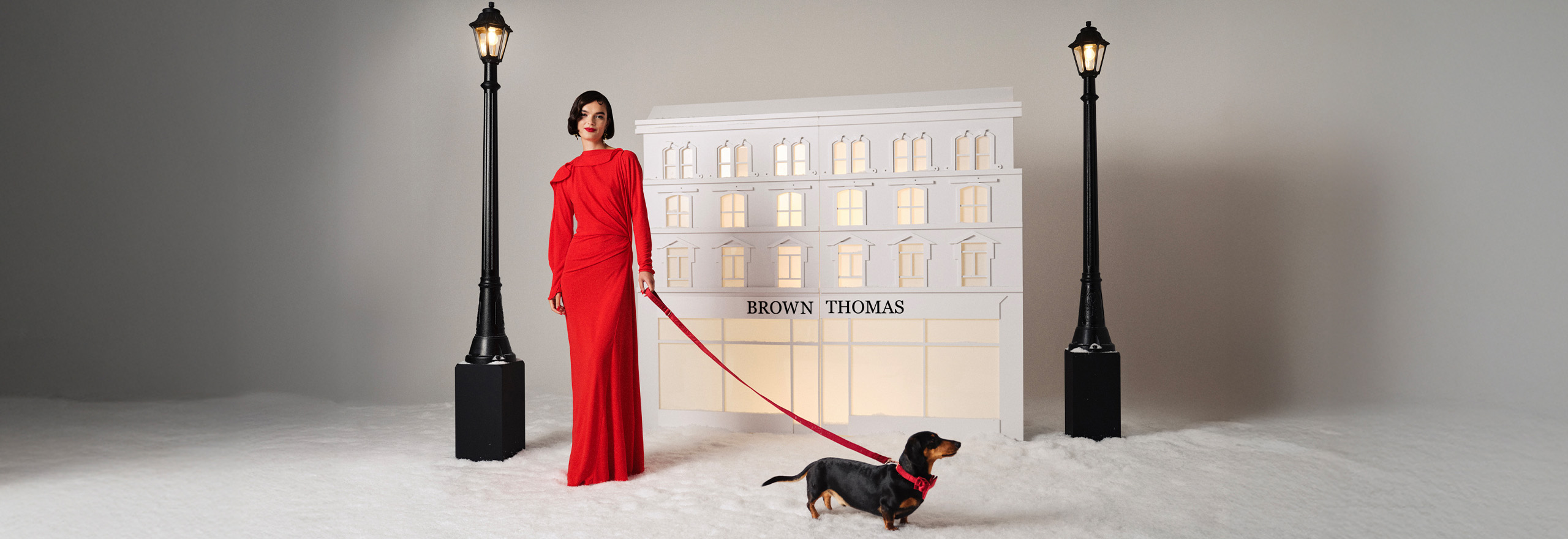 A woman in a red dress holds a dachshund on a leash, standing near a model of a 'Brown Thomas' building between two lampposts.