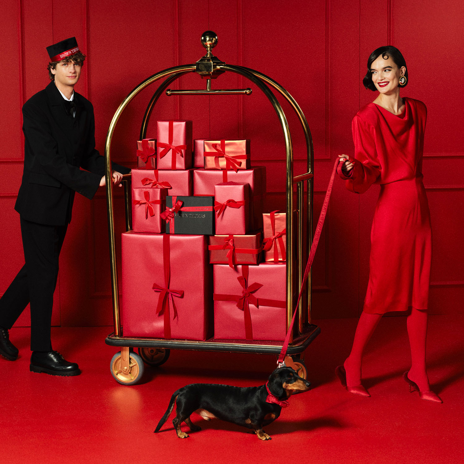 A woman in red holds a dachshund's leash next to a bellhop with a cart full of red and black gift boxes against a red background.