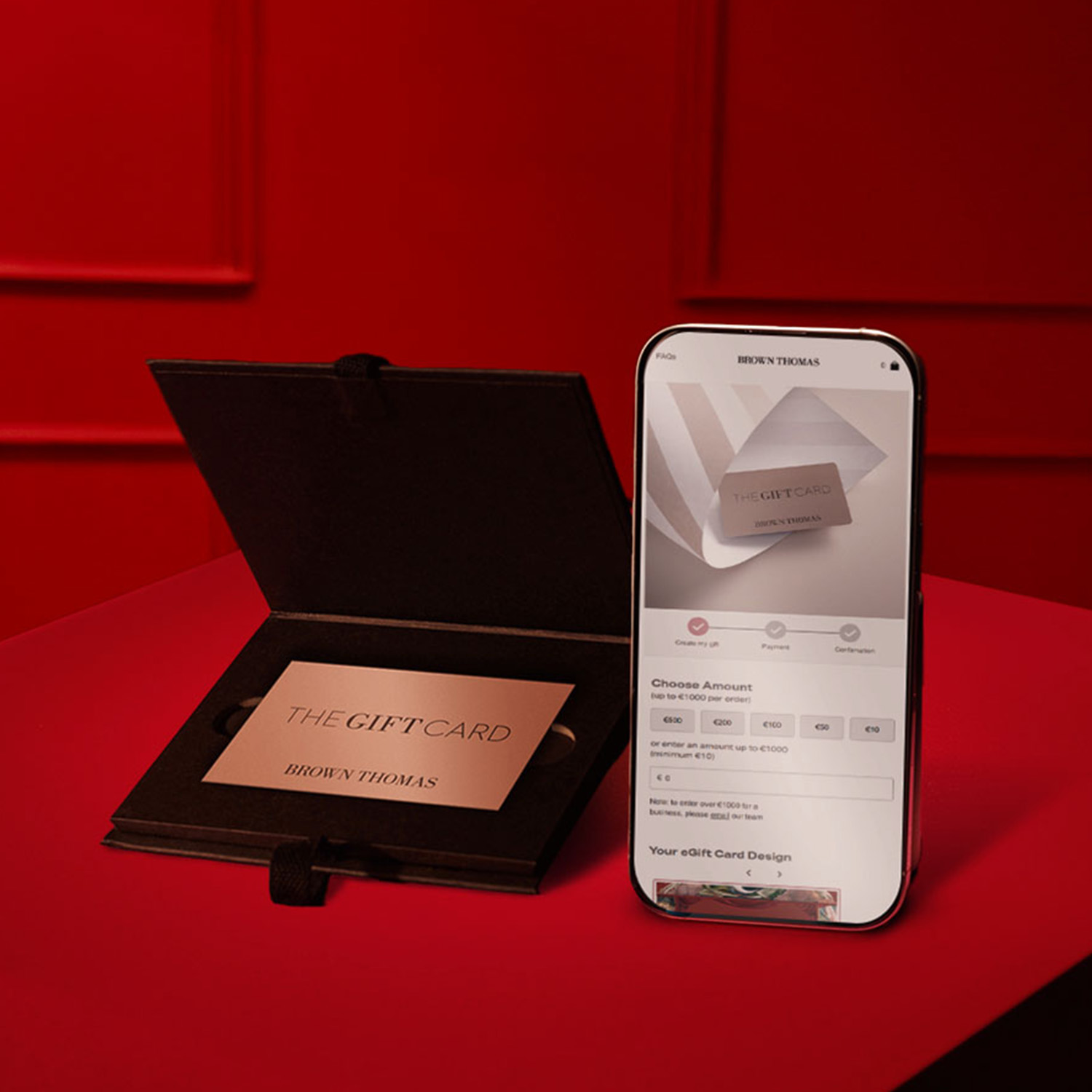 A gift card is displayed in an open black box next to a smartphone showing gift card options against a red background.