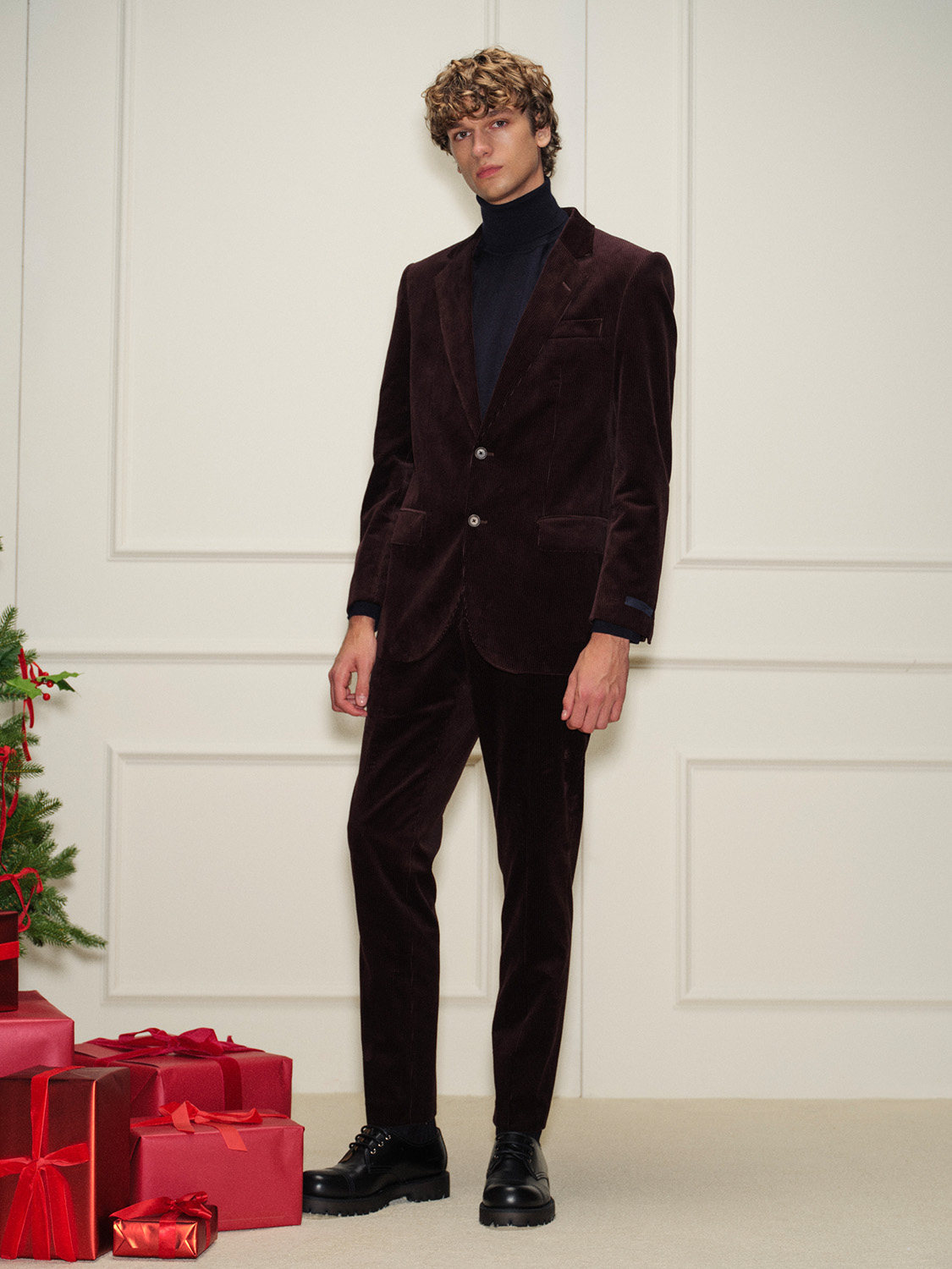 A person in a dark brown velvet suit and black turtleneck stands near a wall, with wrapped gifts and a hint of a Christmas tree nearby.
