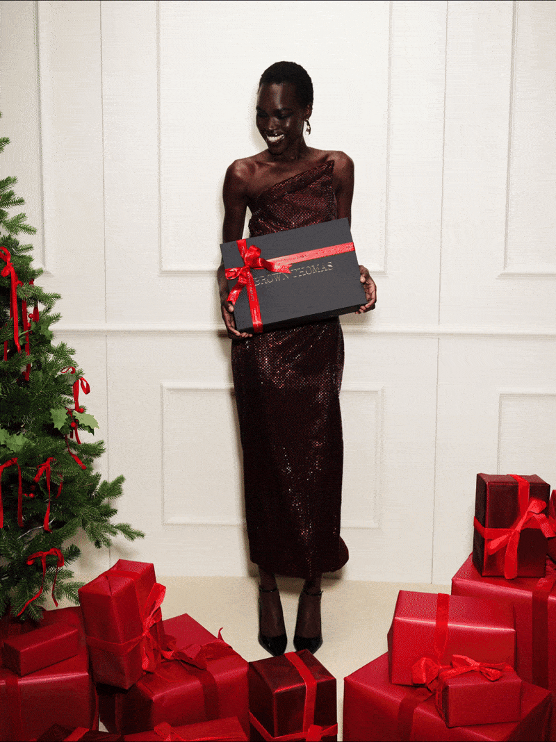A person in a sequined dress holding a gift box stands beside a decorated tree and red presents.