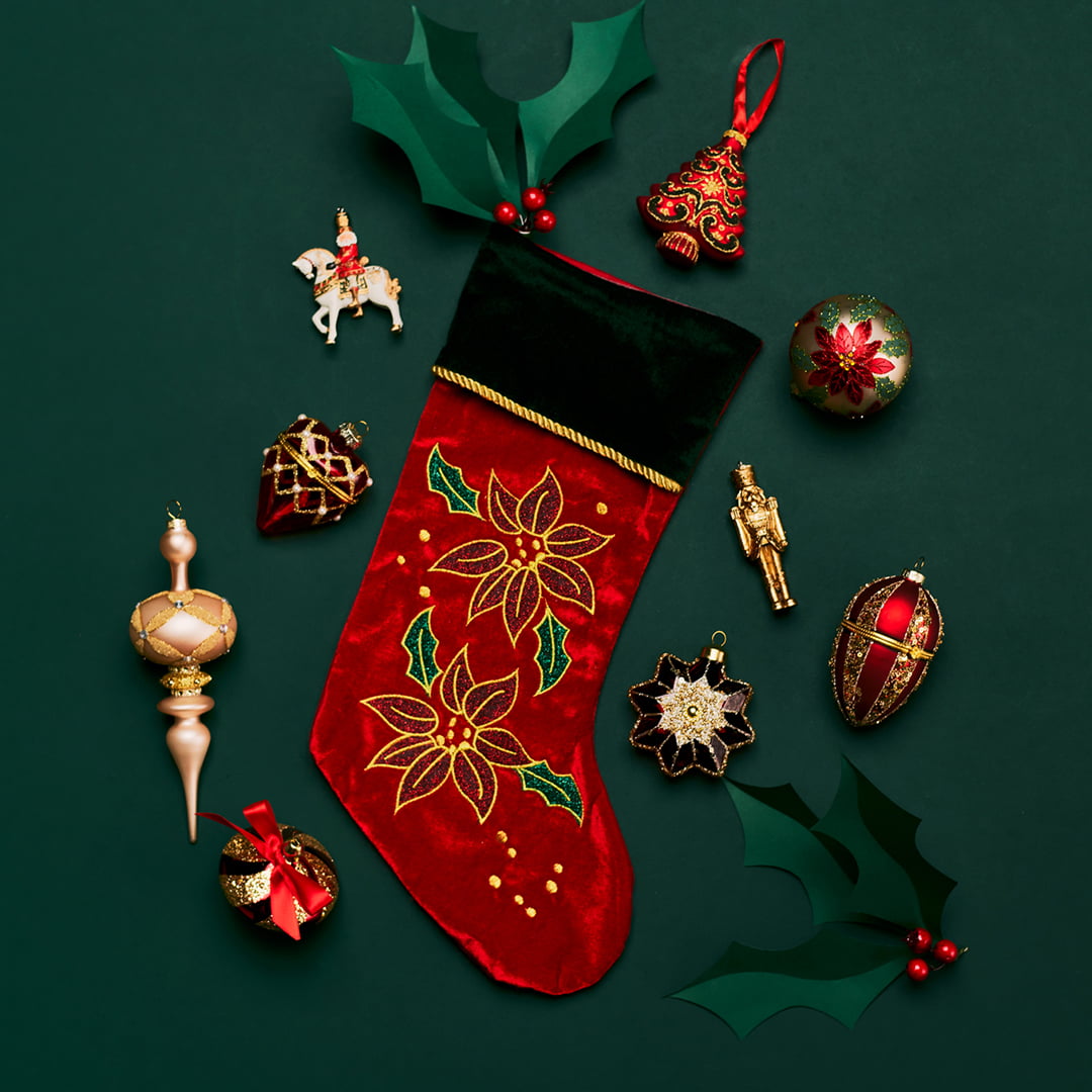 Red Christmas stocking with gold poinsettias, surrounded by festive ornaments and holly on a dark green background.