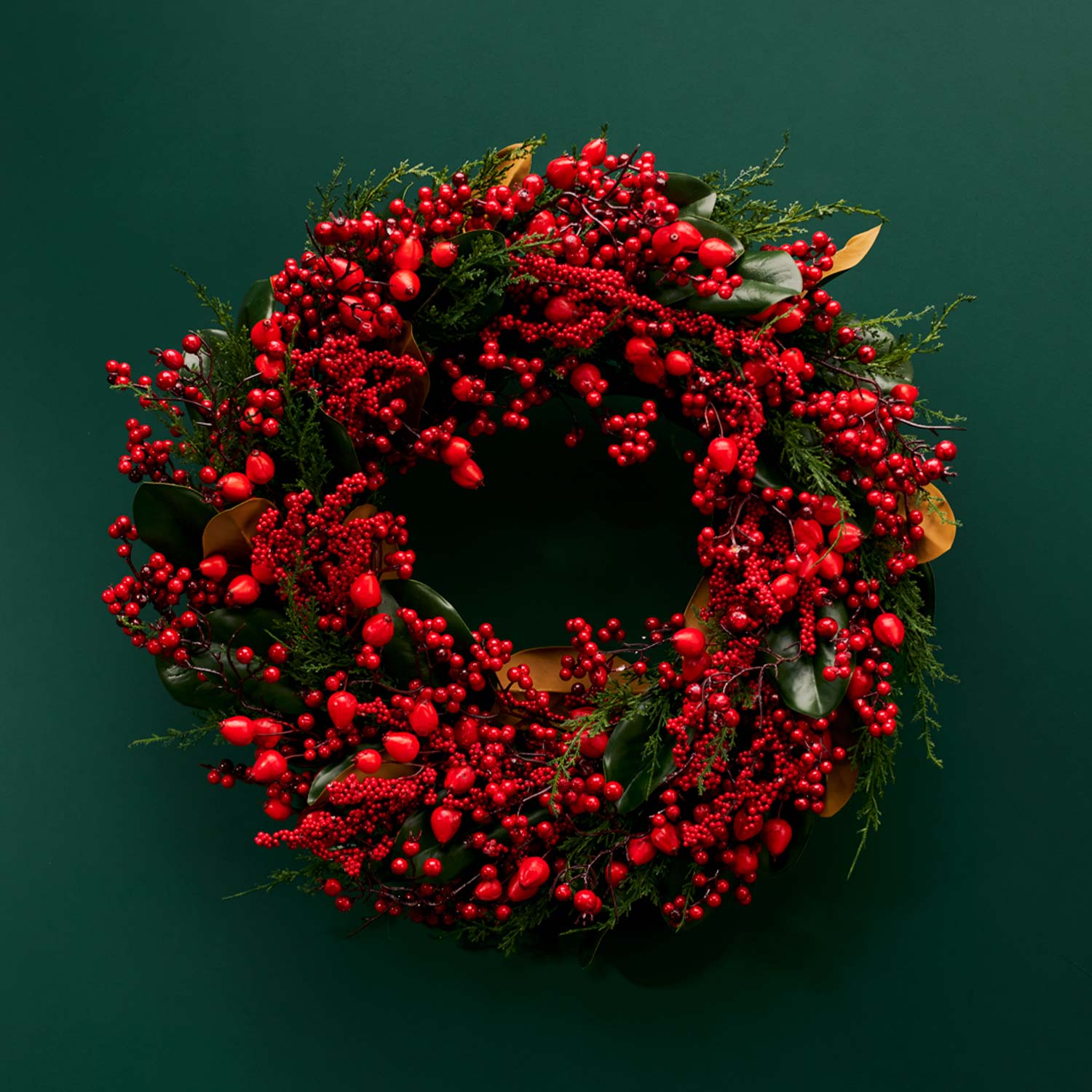 A festive wreath made of red berries and green leaves against a dark green background.