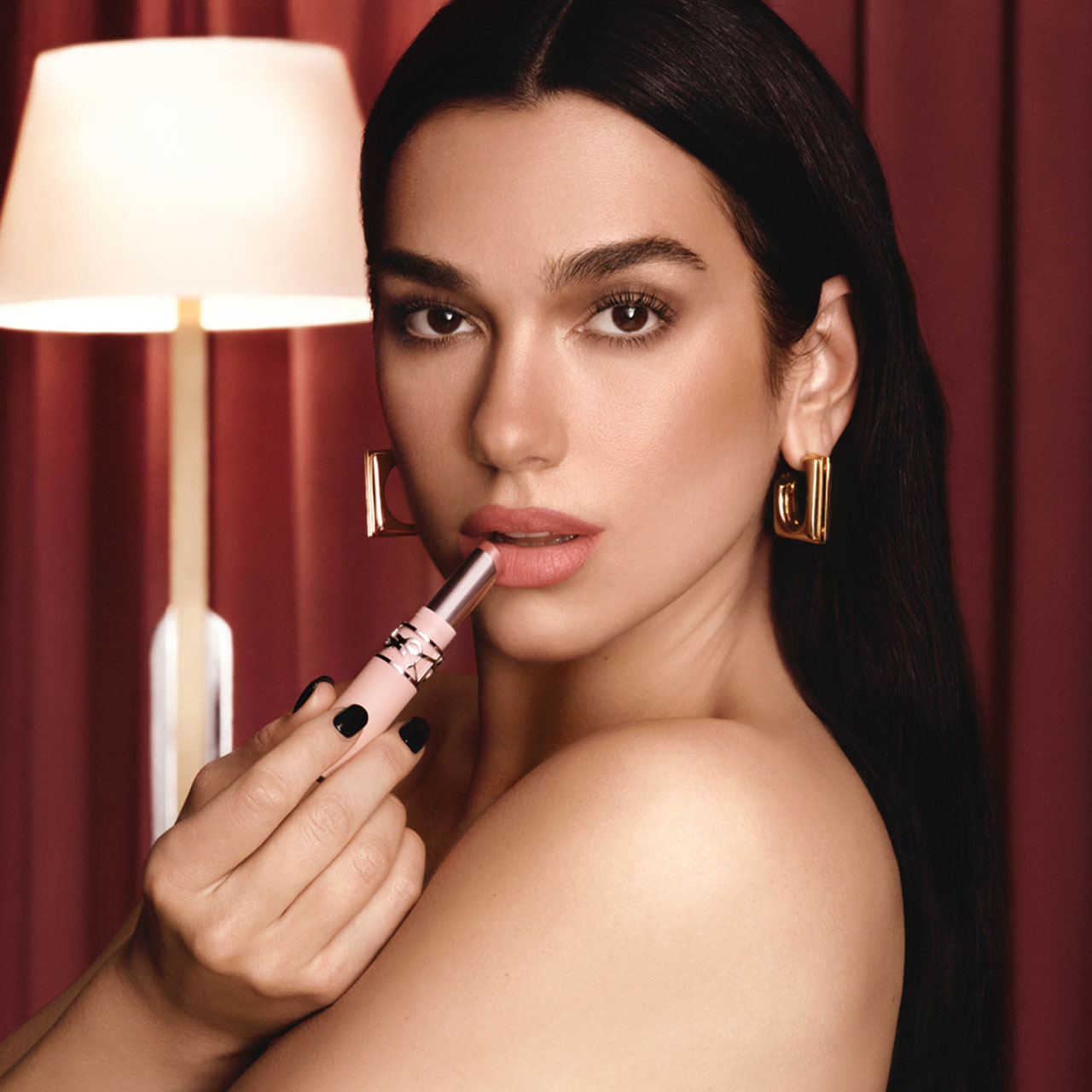 Woman with black long hair applying pink lipstick against burgundy backdrop, wearing gold hoop earrings