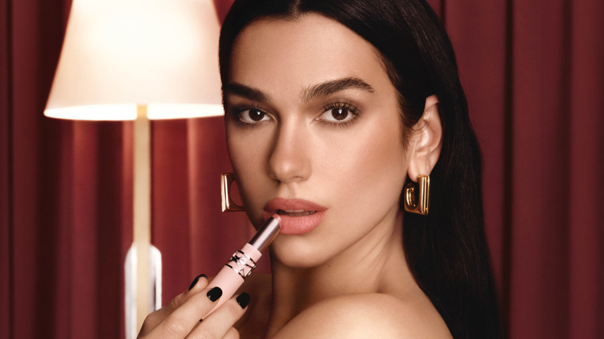 Woman with long dark hair and gold hoop earrings applying pink lip product, against burgundy curtains with table lamp