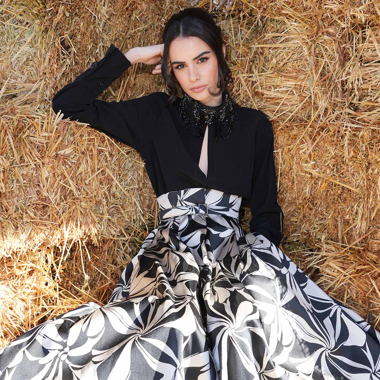 A person in a black top and patterned skirt lies on straw, with a stylish and elegant pose.