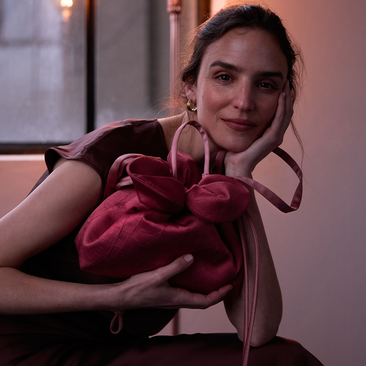 A woman in a burgundy dress holds a matching, tied bag, leaning on her hand, with a soft smile.