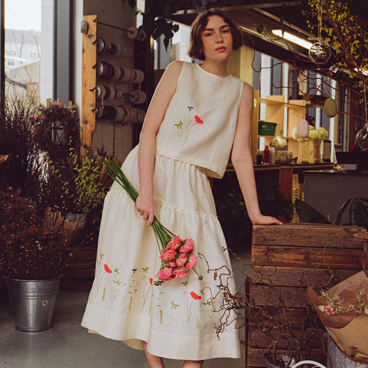 A person in a white floral outfit holds pink flowers, standing in a rustic, plant-filled space.