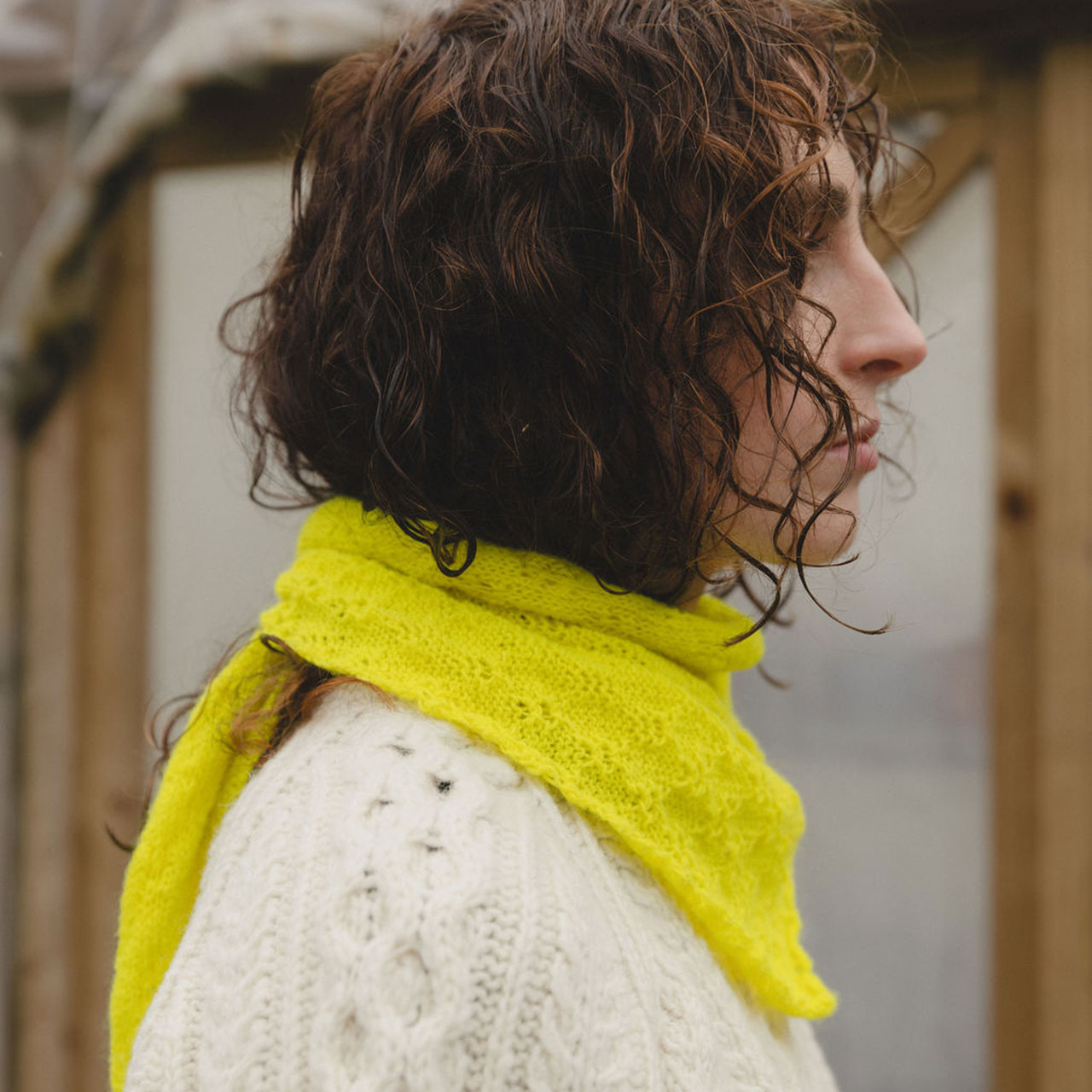 Person with curly hair wears a yellow scarf and white sweater, standing in front of a wooden structure.
