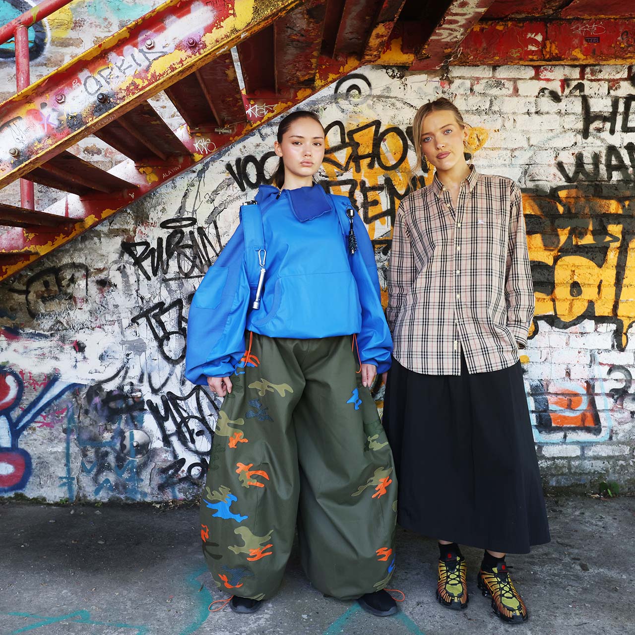 Two models wearing contemporary fashion against a colourful graffitied wall. Left model wears bright blue jacket with olive cargo trousers; right model wears checked shirt with black skirt.