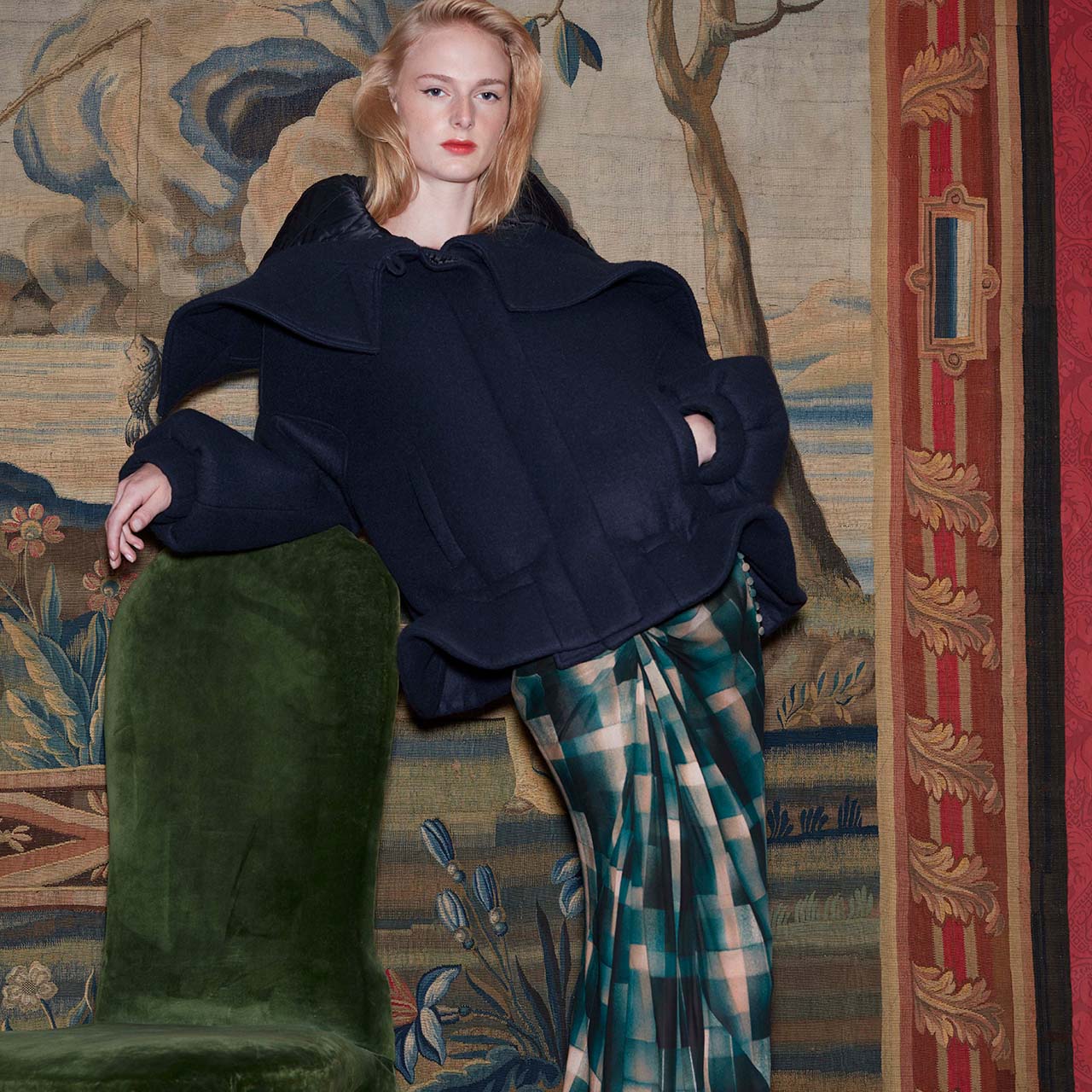 Model wearing full length gown and coat, leaning against a chair, against Victorian style room backdrop