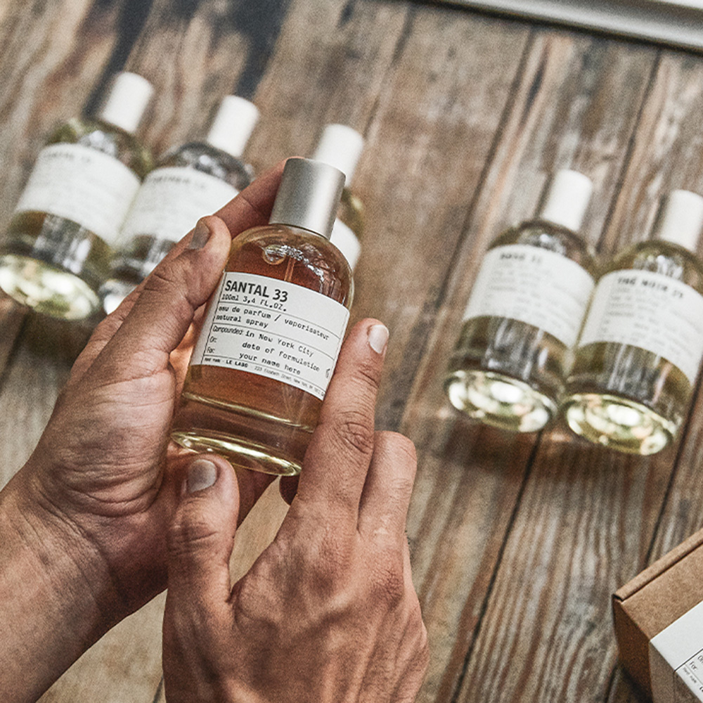 Hand holding Santal 33 fragrance bottle with additional Le Labo bottles displayed on wooden surface