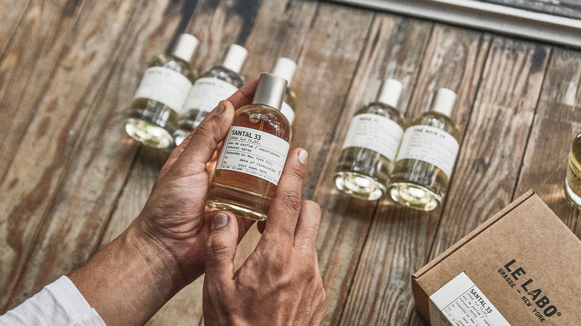 Hand holding Santal 33 fragrance bottle with additional Le Labo perfume bottles displayed on wooden surface
