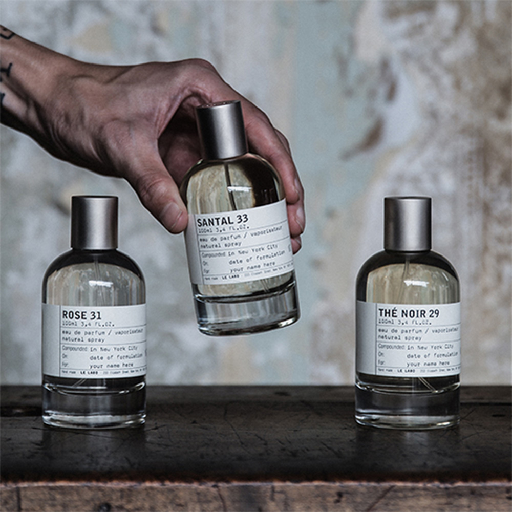 Three unisex Le Labo fragrances displayed: Rose 31, Santal 33, and Thé Noir 29 with hand holding centre bottle