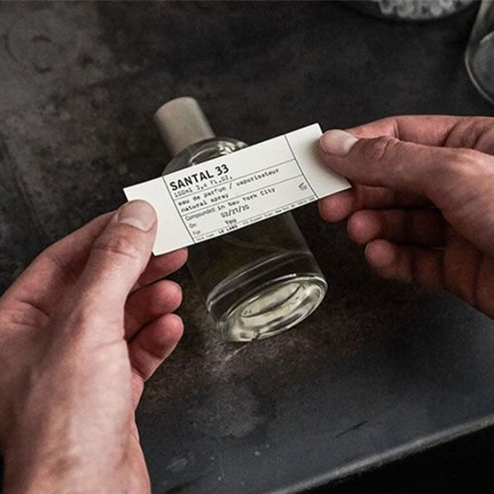 Hands holding Santal 33 fragrance label with bottle visible in background on dark surface